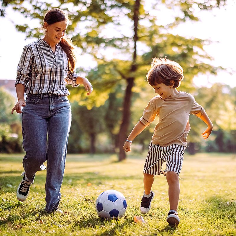 A mum and her son laughing as they kick a football in the park